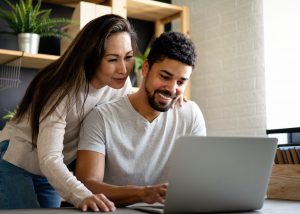 Couple sitting in front of computer where both are smiling and are observing computer.  Woman is looking over mans shoulder. 