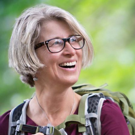 Middle aged woman wearing glasses and a backpack smiling as she is hiking outside.